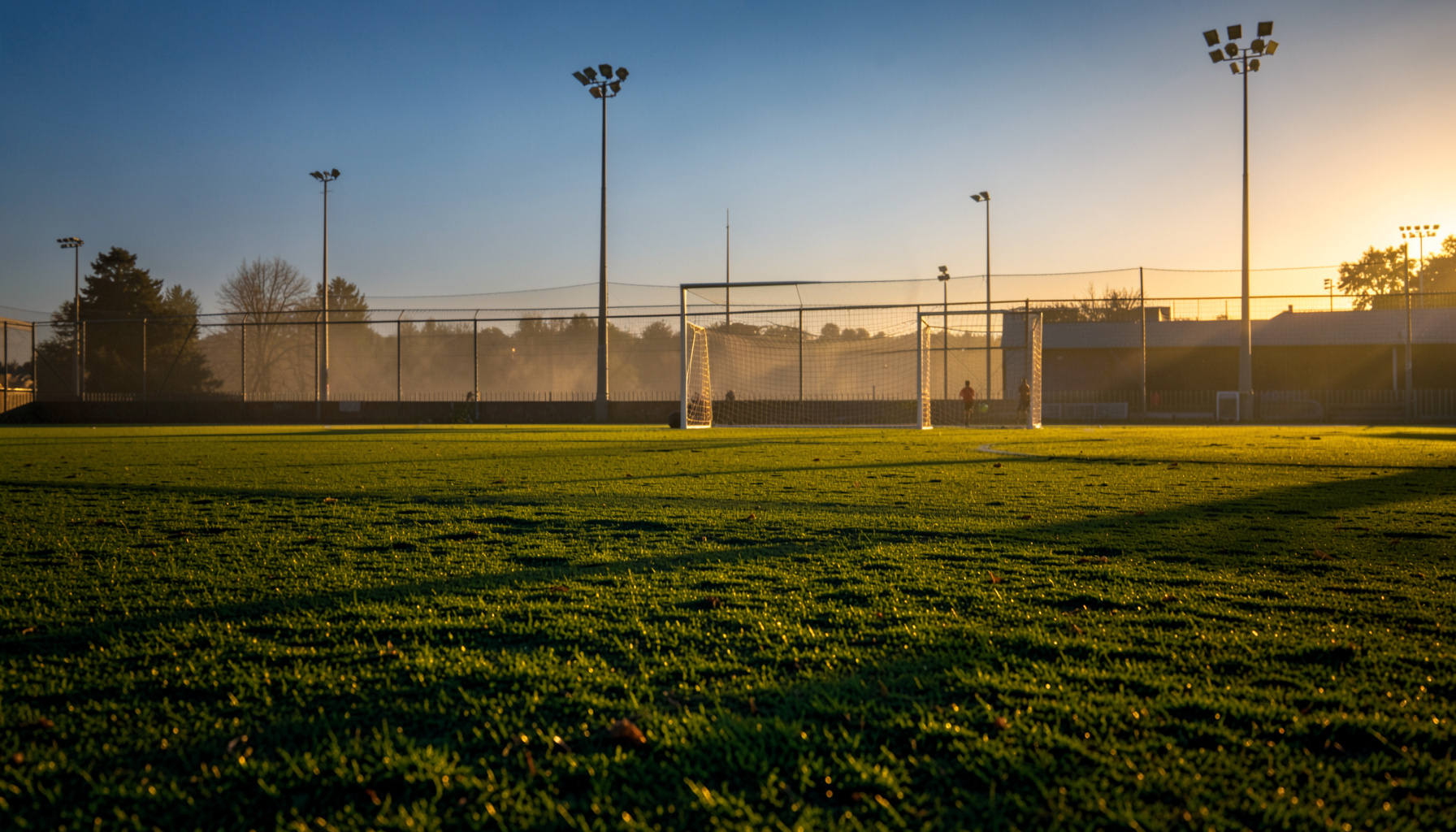 Campo de futebol 7 ao entardecer, com relva verde intensa iluminada por luz dourada e balizas ao fundo, atmosfera de competição nacional