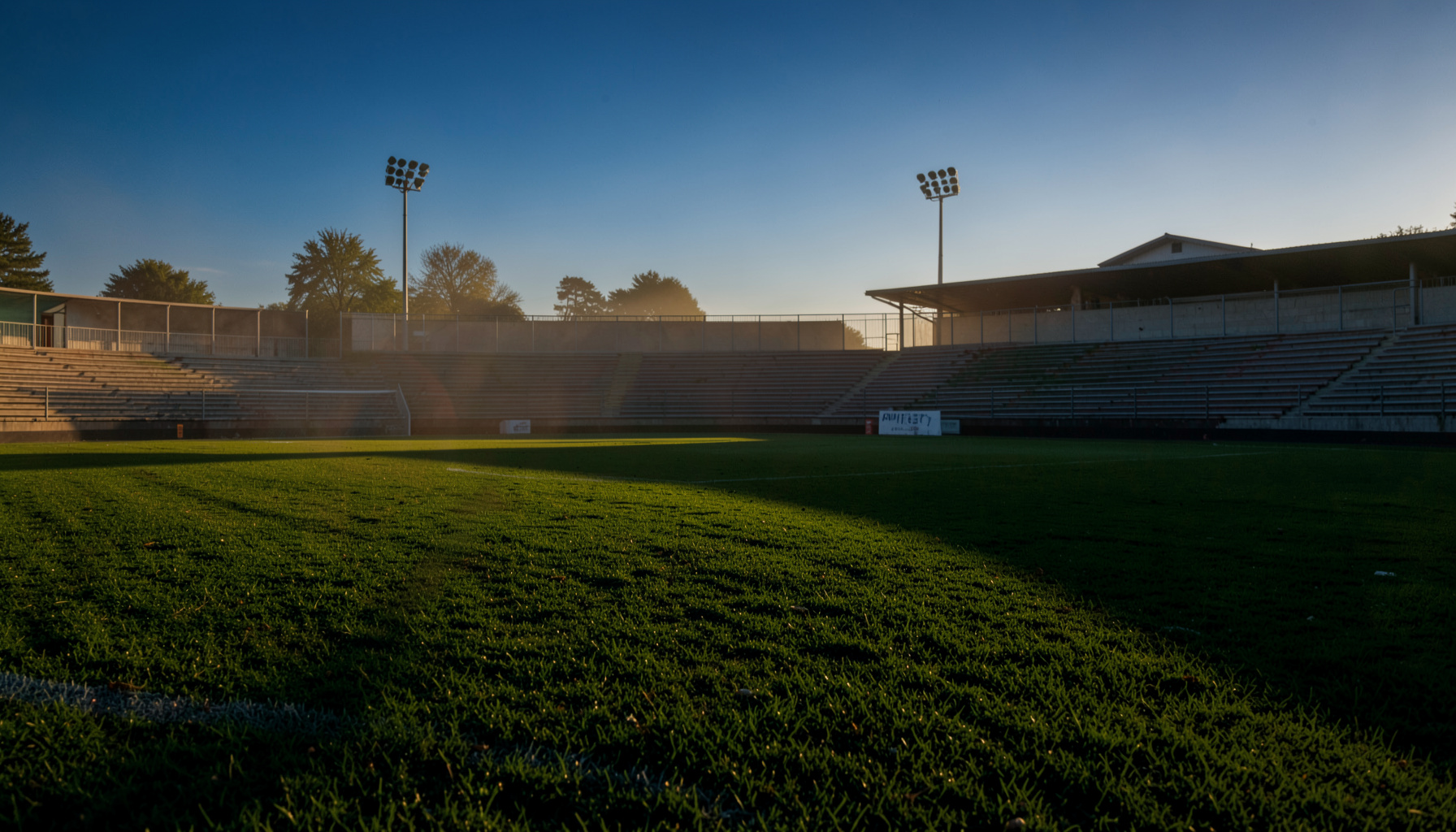 Campo de futebol 7 ao entardecer com relva verde intensa e iluminação artificial suave, bancada vazia ao fundo
